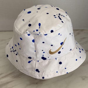 May include: A white bucket hat featuring a blue paint splatter pattern across the crown and brim. The hat includes an embroidered gold Nike swoosh logo on the front panel, positioned above the brim, with a rounded top and a downward sloping brim.