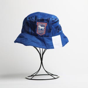 May include: A blue bucket hat with a white stripe and a team logo patch. The patch features a white horse and the words "IPSWICH TOWN" in red and white. The hat is displayed on a black metal stand.