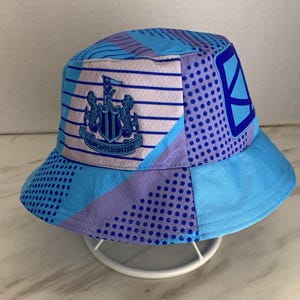 May include: A bucket hat featuring a light blue and purple geometric pattern with a mix of stripes and dots. The front displays the Newcastle United football club crest in blue and white, along with a dark blue graphic design on the side panel.