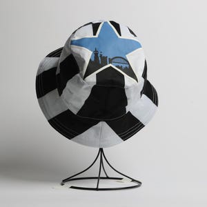 May include: A black and white bucket hat with a star design and a silhouette of a city skyline. The hat is displayed on a black metal stand against a white background. The hat has a wide brim.