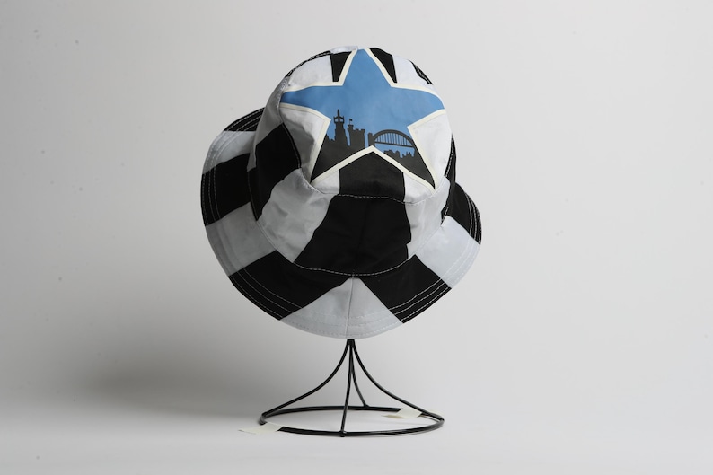 May include: A black and white bucket hat with a star design and a silhouette of a city skyline. The hat is displayed on a black metal stand against a white background. The hat has a wide brim.