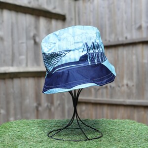 May include: A blue bucket hat with a bridge design. The hat features a light blue crown with a dark blue band and a bridge graphic. The hat is displayed on a black metal stand against a wooden fence.