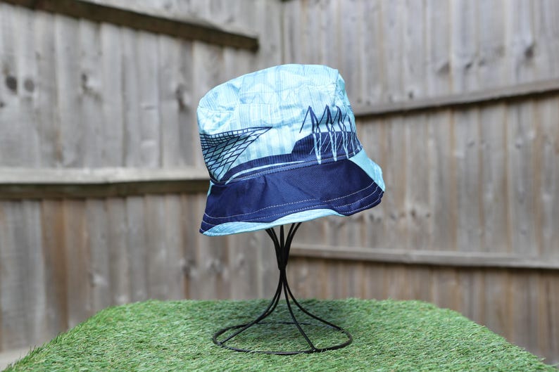 May include: A blue bucket hat with a bridge design. The hat features a light blue crown with a dark blue band and a bridge graphic. The hat is displayed on a black metal stand against a wooden fence.