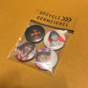 Puede incluir: Un conjunto de cuatro botones redondos con imágenes de un futbolista. Los botones están empaquetados en una bolsa de plástico transparente con una etiqueta de papel marrón que dice "UPCYCLE SCHMEICHEL". Los botones tienen un acabado brillante.