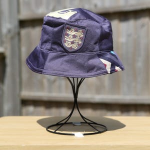 May include: A navy blue bucket hat with a white and cream design on the top and a patch featuring the England national team emblem. The hat is displayed on a black metal stand.