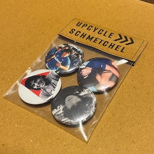 Op de afbeelding: Een set van vier ronde buttons in een doorzichtige plastic verpakking. De buttons tonen afbeeldingen van een persoon in verschillende poses en een teamfoto. De verpakking heeft een bruin papieren label met de tekst "UPCYCLE SCHMETCHEL".