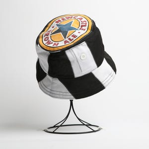 May include: Bucket hat in black and white chequered pattern, featuring a Newcastle Brown Ale logo. The logo is a yellow circle with a blue star and the words "Newcastle Brown Ale". The hat has a button detail and is displayed on a black stand.