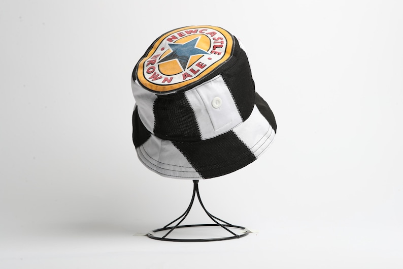 May include: Bucket hat in black and white chequered pattern, featuring a Newcastle Brown Ale logo. The logo is a yellow circle with a blue star and the words "Newcastle Brown Ale". The hat has a button detail and is displayed on a black stand.