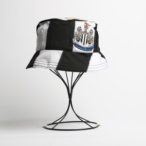 May include: A black and white chequered bucket hat, featuring the Newcastle United crest. The hat is displayed on a black metal stand. The hat has a patch with the team's emblem.
