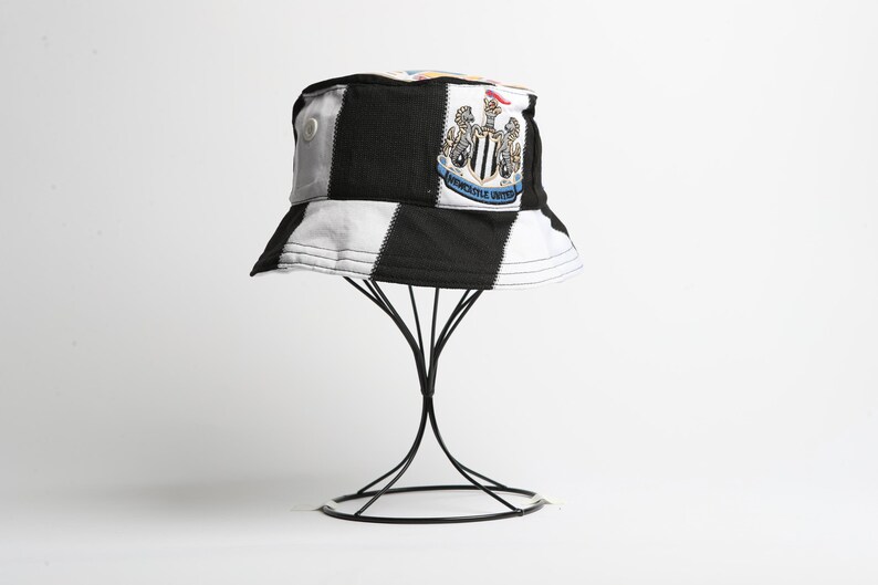May include: A black and white chequered bucket hat, featuring the Newcastle United crest. The hat is displayed on a black metal stand. The hat has a patch with the team's emblem.