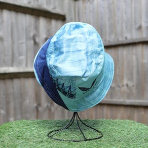 May include: A blue and navy bucket hat with a graphic design and logo. The hat is displayed on a black metal stand against a wooden fence and green surface.