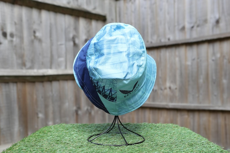 May include: A blue and navy bucket hat with a graphic design and logo. The hat is displayed on a black metal stand against a wooden fence and green surface.