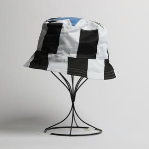May include: A black and white striped bucket hat with a light blue interior. The hat is displayed on a black wire stand against a white background. The hat's design features bold, vertical stripes.