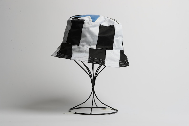 May include: A black and white striped bucket hat with a light blue interior. The hat is displayed on a black wire stand against a white background. The hat's design features bold, vertical stripes.