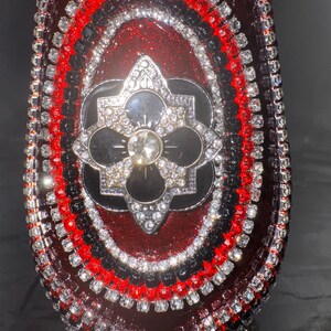 May include: Close-up of a decorative, ornate object with a deep red, glittery surface. The object is embellished with rows of sparkling rhinestones in red, black, and silver. A central, multi-layered design features a flower-like shape.
