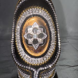 May include: A black decorative bottle with intricate rhinestone and gold embellishments. The bottle features an oval design with a central floral motif, surrounded by layers of sparkling rhinestones and gold accents, creating a luxurious appearance.