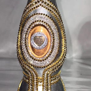 May include: A decorative bottle with a silver metallic finish, adorned with gold and silver rhinestones. The bottle features a crown-shaped top and an oval centerpiece with text. The design includes intricate patterns and embellishments.