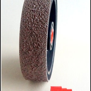 May include: A large, textured, brown abrasive disc with a black interior and a red center. A small, red cylindrical component is positioned below the disc. The disc appears to be for grinding or sanding.