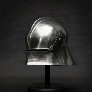 K&ouml;nnte beinhalten: Ein polierter, silberfarbener Helm mit abgerundeter Oberseite und Visier. Der Helm steht auf einem schwarzen St&auml;nder vor einem dunklen Hintergrund. Die Oberfl&auml;che des Helms reflektiert das Licht und hebt seine Form hervor.