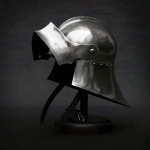 K&ouml;nnte beinhalten: Ein polierter Stahlhelm mit Visier, ausgestellt auf einem schwarzen St&auml;nder. Der Helm hat eine abgerundete Oberseite und einen ausgestellten unteren Teil mit Nieten am Rand. Lederriemen sind befestigt.