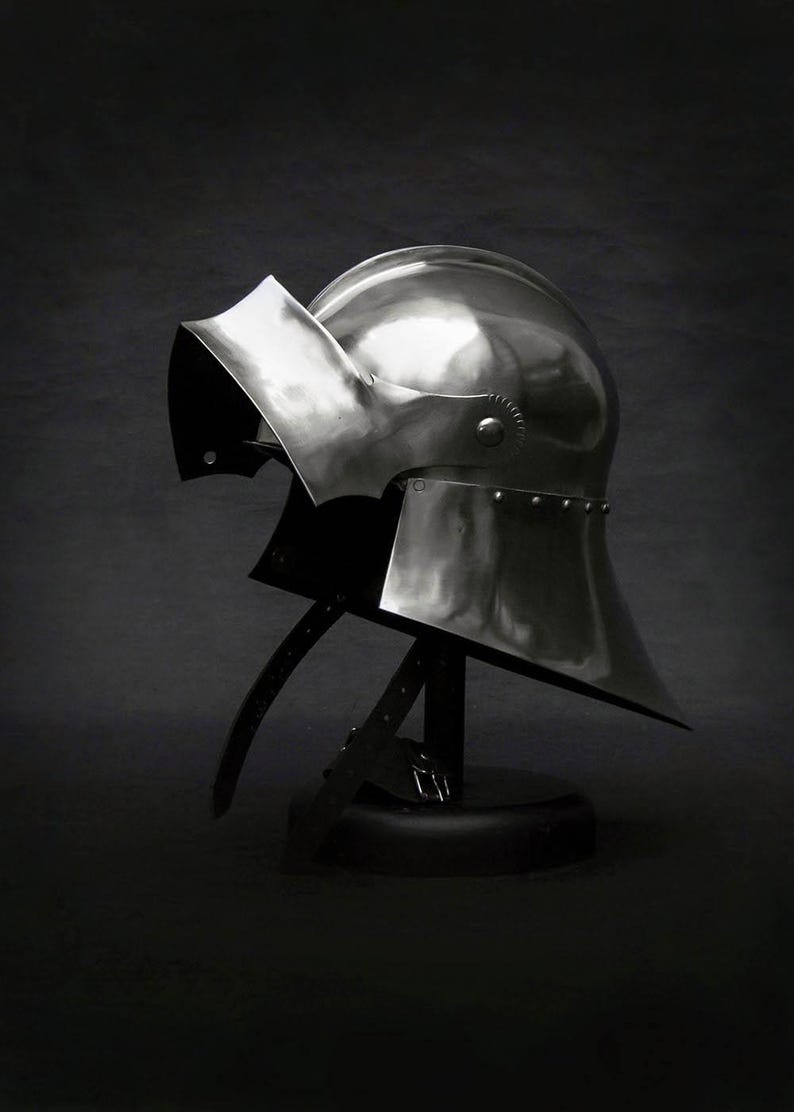 K&ouml;nnte beinhalten: Ein polierter Stahlhelm mit Visier, ausgestellt auf einem schwarzen St&auml;nder. Der Helm hat eine abgerundete Oberseite und einen ausgestellten unteren Teil mit Nieten am Rand. Lederriemen sind befestigt.