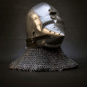 May include: A polished steel knight's helmet with a pointed visor and numerous ventilation holes. The helmet features a decorative element on top and is connected to a chainmail neck guard. A leather strap secures the helmet.