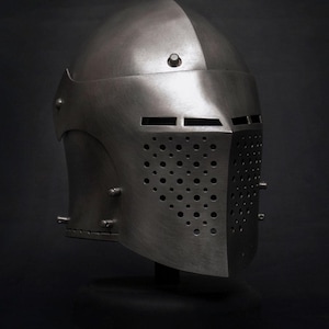 Op de afbeelding: Een zilverkleurige middeleeuwse helm met een afgeronde bovenkant en een vizier met talrijke kleine gaatjes en horizontale sleuven. De helm wordt gepresenteerd tegen een donkere achtergrond.