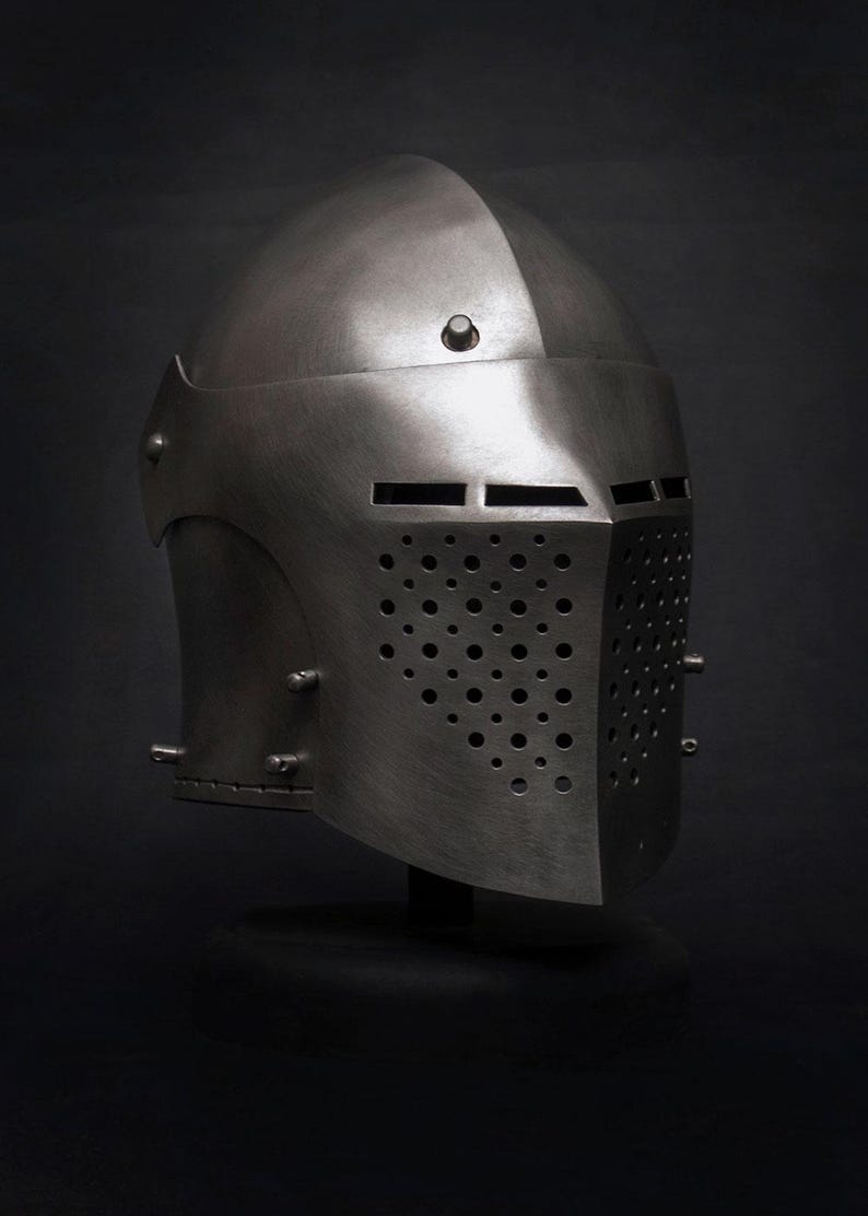 Op de afbeelding: Een zilverkleurige middeleeuwse helm met een afgeronde bovenkant en een vizier met talrijke kleine gaatjes en horizontale sleuven. De helm wordt gepresenteerd tegen een donkere achtergrond.
