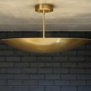 Op de afbeelding: Een goudkleurige, komvormige plafondlamp. Het licht wordt opgehangen aan een bijpassende goudkleurige plafondbevestiging door een slanke staaf. De armatuur staat tegen een donkere bakstenen muur.