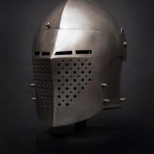 Op de afbeelding: Een zilverkleurige middeleeuwse helm met een afgeronde bovenkant en een frontplaat met talrijke kleine gaatjes en horizontale sleuven voor zicht en ventilatie. De helm wordt op een zwarte standaard tegen een donkere achtergrond gepresenteerd.