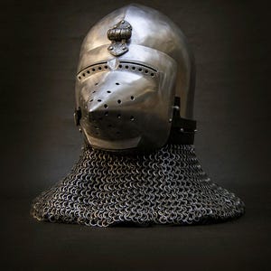 May include: A polished metal helmet with a visor and a chainmail neck guard. The helmet has a rounded top and a pointed visor with several small holes. The chainmail extends down to the shoulders. The helmet is likely from the medieval period.