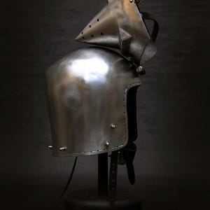 May include: A polished steel helmet with a pointed visor and riveted construction. The medieval-style helmet is displayed on a black stand against a dark background. The helmet's design includes a neck guard and a hinged visor.