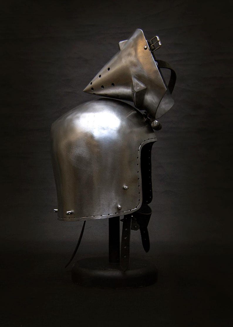 May include: A polished steel helmet with a pointed visor and riveted construction. The medieval-style helmet is displayed on a black stand against a dark background. The helmet's design includes a neck guard and a hinged visor.