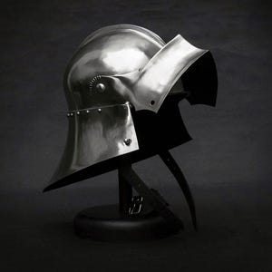 K&ouml;nnte beinhalten: Ein polierter Stahlhelm, vermutlich aus dem Mittelalter, wird auf einem schwarzen St&auml;nder pr&auml;sentiert. Der Helm hat eine abgerundete Oberseite, ein Visier und einen Nackenschutz. Nieten und Riemen sind sichtbar. Der Hintergrund ist dunkel und einfarbig.