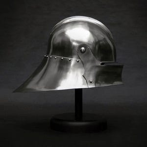 K&ouml;nnte beinhalten: Ein polierter, silberfarbener mittelalterlicher Helm mit breitem, nach unten geneigtem Visier. Der Helm ist auf einem schwarzen St&auml;nder montiert, der seine reflektierende Oberfl&auml;che und die Nietdetails zeigt. Historisches R&uuml;stungsdesign.