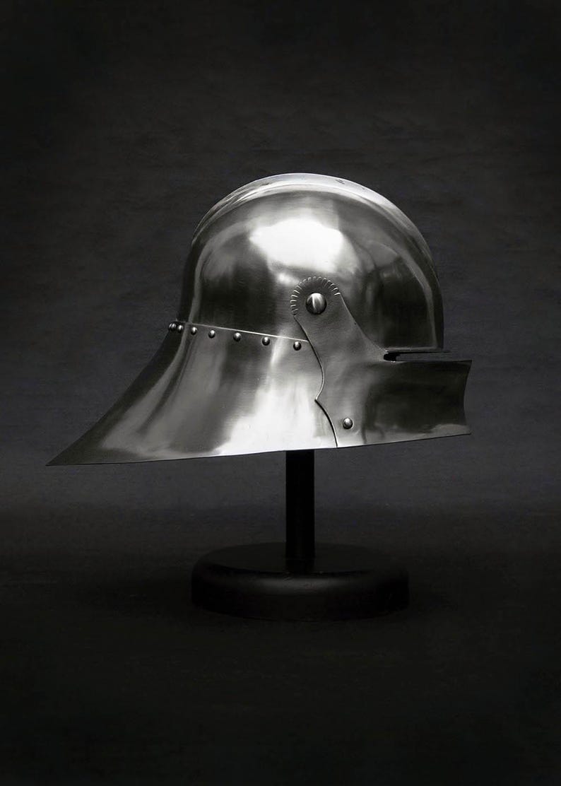 K&ouml;nnte beinhalten: Ein polierter, silberfarbener mittelalterlicher Helm mit breitem, nach unten geneigtem Visier. Der Helm ist auf einem schwarzen St&auml;nder montiert, der seine reflektierende Oberfl&auml;che und die Nietdetails zeigt. Historisches R&uuml;stungsdesign.
