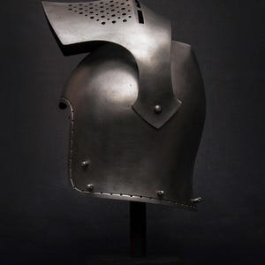 Op de afbeelding: Een middeleeuwse stalen helm met vizier en nekbescherming, gepresenteerd op een zwarte standaard tegen een donkere achtergrond. De helm heeft een gebogen ontwerp met klinknagels en kleine ventilatiegaten op het vizier.