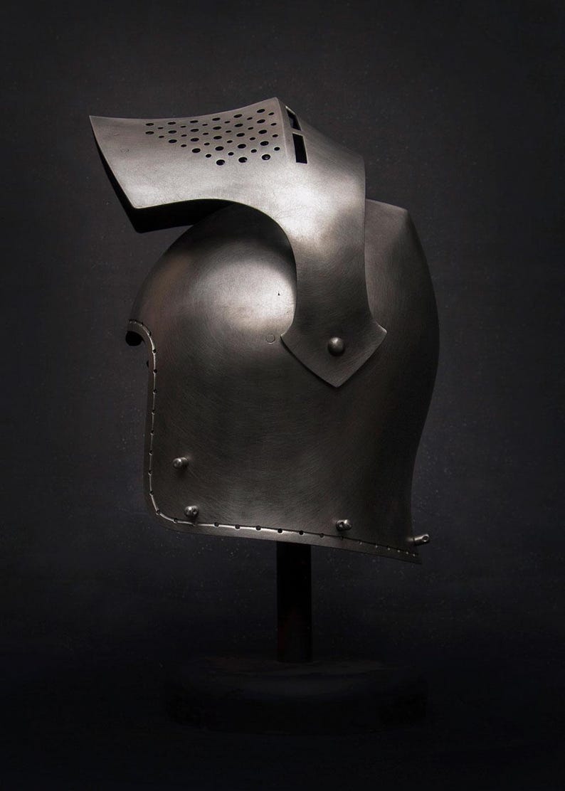 Op de afbeelding: Een middeleeuwse stalen helm met vizier en nekbescherming, gepresenteerd op een zwarte standaard tegen een donkere achtergrond. De helm heeft een gebogen ontwerp met klinknagels en kleine ventilatiegaten op het vizier.