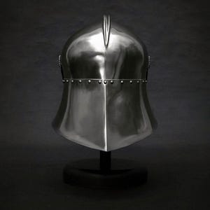 K&ouml;nnte beinhalten: Ein polierter, silberfarbener mittelalterlicher Helm mit spitzer Oberseite und genieteten Zierleisten. Der Helm wird auf einem schwarzen St&auml;nder vor dunklem Hintergrund pr&auml;sentiert und zeigt seine reflektierende Oberfl&auml;che und sein historisches Design.