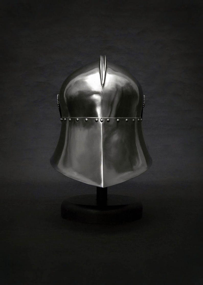K&ouml;nnte beinhalten: Ein polierter, silberfarbener mittelalterlicher Helm mit spitzer Oberseite und genieteten Zierleisten. Der Helm wird auf einem schwarzen St&auml;nder vor dunklem Hintergrund pr&auml;sentiert und zeigt seine reflektierende Oberfl&auml;che und sein historisches Design.