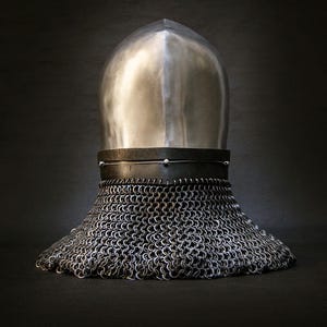May include: A medieval helmet with a polished, silver-colored top and a chainmail neck guard. The helmet sits on a dark surface, with the chainmail extending outward. The design is reminiscent of historical armor.