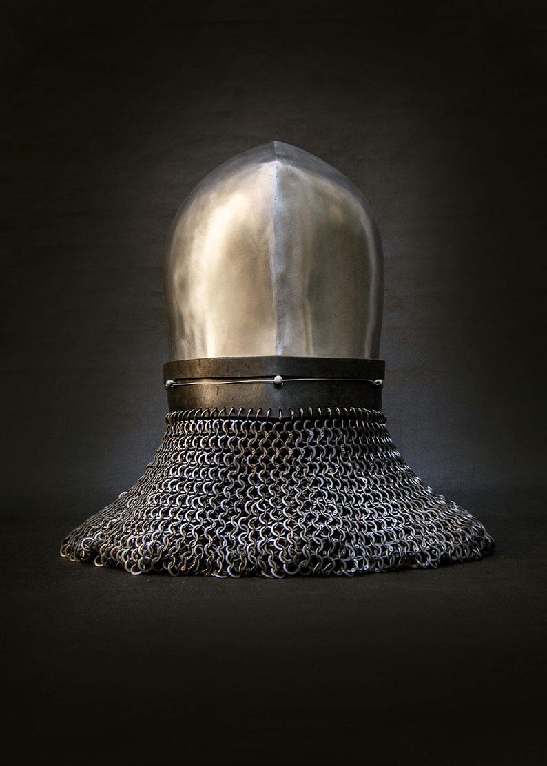 May include: A medieval helmet with a polished, silver-colored top and a chainmail neck guard. The helmet sits on a dark surface, with the chainmail extending outward. The design is reminiscent of historical armor.