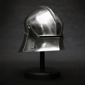 K&ouml;nnte beinhalten: Ein polierter, silberfarbener mittelalterlicher Helm, der auf einem schwarzen St&auml;nder pr&auml;sentiert wird. Der Helm hat eine abgerundete Oberseite mit einem markanten Mittelgrat und einem ausgestellten unteren Rand. Nieten und dekorative Details sind sichtbar.