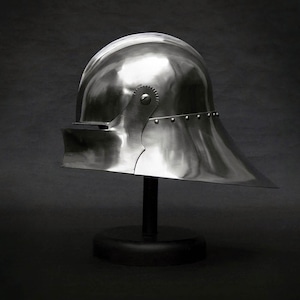 K&ouml;nnte beinhalten: Ein polierter, silberfarbener mittelalterlicher Helm mit rundem Oberteil und Visier. Der Helm steht auf einem schwarzen St&auml;nder vor dunklem Hintergrund. Das Visier ragt nach au&szlig;en, und der Helm ist seitlich genietet.