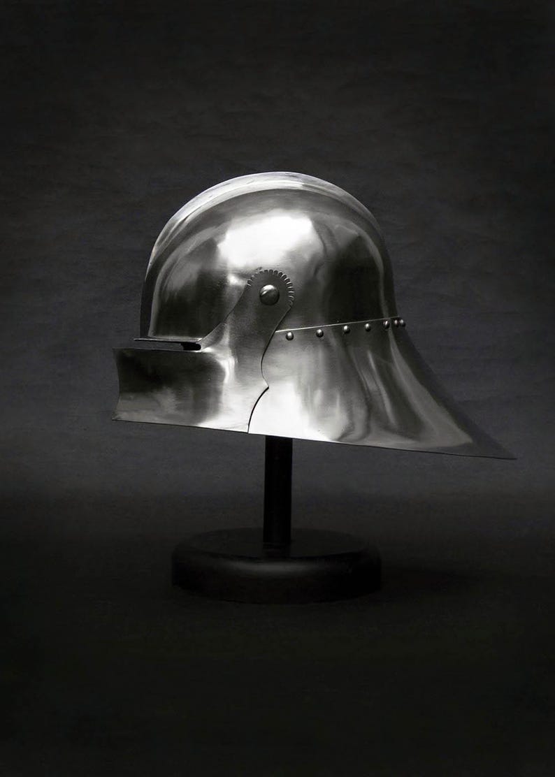 K&ouml;nnte beinhalten: Ein polierter, silberfarbener mittelalterlicher Helm mit rundem Oberteil und Visier. Der Helm steht auf einem schwarzen St&auml;nder vor dunklem Hintergrund. Das Visier ragt nach au&szlig;en, und der Helm ist seitlich genietet.