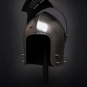 Op de afbeelding: Een donker metalen helm met een gebogen vizier en een afgeronde bovenkant. De helm heeft klinknagels langs de randen en wordt tentoongesteld op een zwarte standaard tegen een donkere achtergrond. Het vizier heeft kleine perforaties.