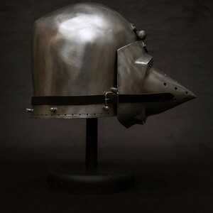 May include: A polished metal helmet with a pointed visor and a black strap. The helmet is displayed on a black stand against a dark background. The helmet's design suggests medieval or historical armor.