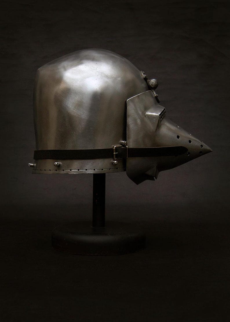 May include: A polished metal helmet with a pointed visor and a black strap. The helmet is displayed on a black stand against a dark background. The helmet's design suggests medieval or historical armor.