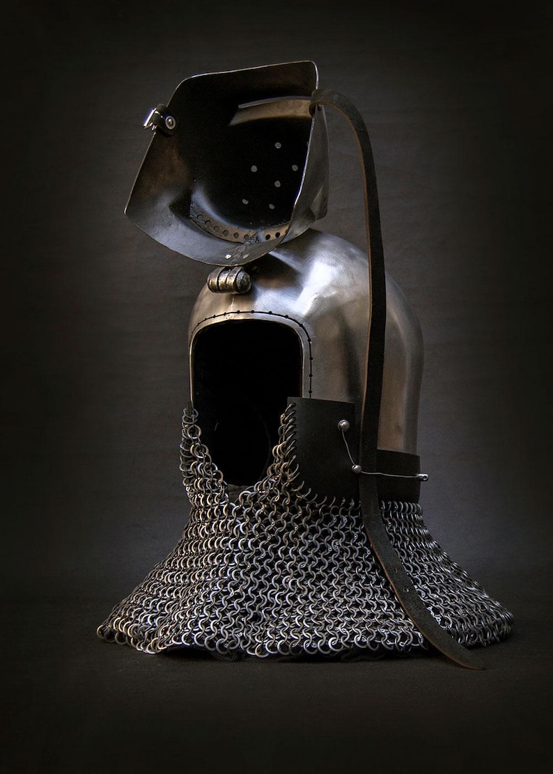 May include: A medieval-style helmet with a hinged visor and chainmail neck guard. The helmet is made of polished metal, with a leather strap and rivets. The visor has several small holes. The overall design suggests historical armor.