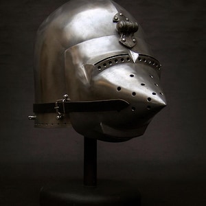 May include: A polished steel medieval helmet with a pointed visor and numerous ventilation holes. The helmet features a decorative element at the crown and a leather strap with a buckle. It is displayed on a black stand against a dark background.